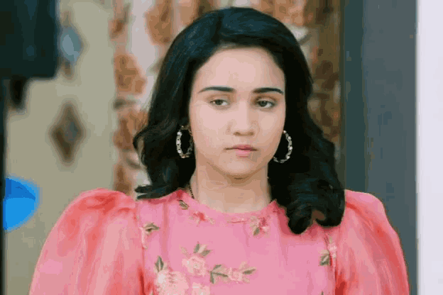 a woman wearing a pink dress and hoop earrings looks serious
