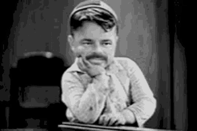 a black and white photo of a man with a mustache sitting at a table with his hand on his chin .