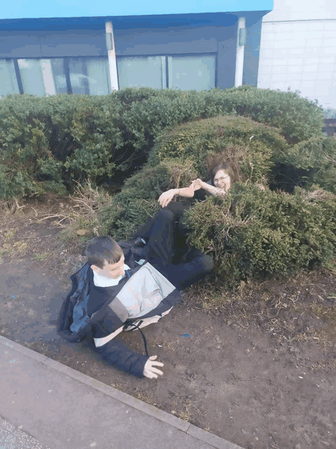 a boy and a girl laying in a bush with a blue building in the background