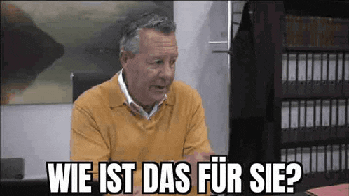 a man in a yellow sweater is sitting at a desk and talking in a german language .