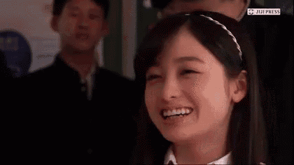 a girl is smiling with her hands folded in front of her face while a man stands behind her .