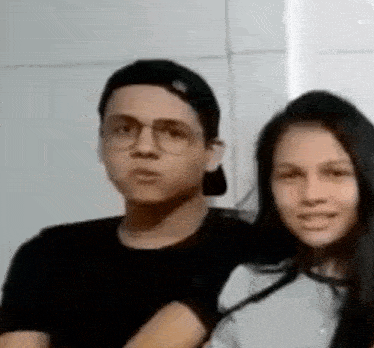 a man and a woman are posing for a picture together . the man is wearing glasses and a baseball cap .