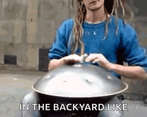 a man in a blue sweater is playing a handpan with the words in the backyard like above him .