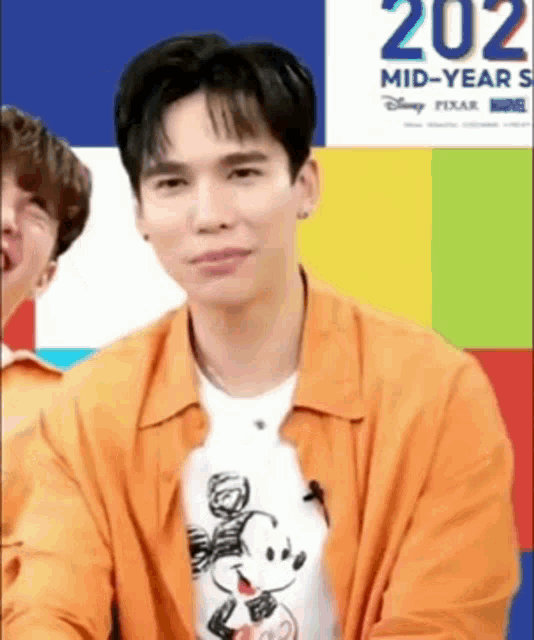 a man wearing an orange jacket and a mickey mouse shirt is sitting in front of a colorful background .