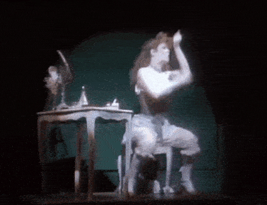 a woman is sitting on a stool on a stage in front of a table and mirror .