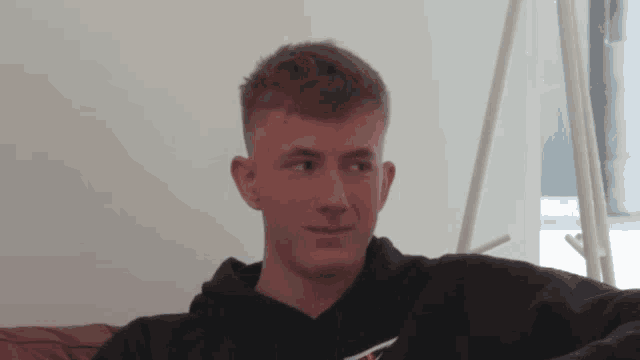 a young man is sitting on a couch wearing a black tommy jeans hoodie