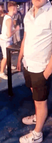 a man in a white shirt and black shorts is standing with his hands in his pockets in front of a crowd of people .