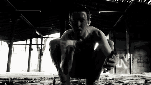 a shirtless man is squatting down in a dark room with graffiti on the wall that says n