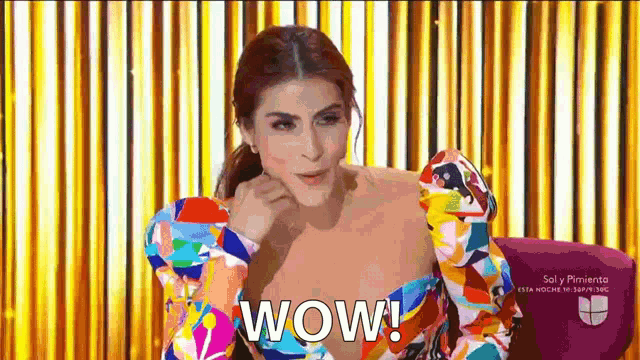 a woman in a colorful dress is sitting in front of a yellow curtain and says wow .