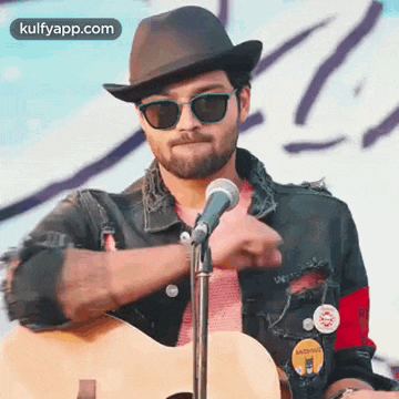 a man wearing a hat , sunglasses and a guitar is playing a guitar in front of a microphone .