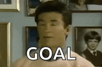 a man in a pink sweater and bow tie is standing in front of a wall with pictures and the word goal written on it .