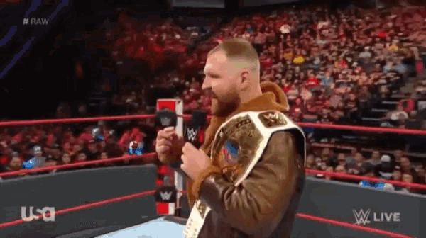 a man in a brown jacket is holding a microphone in a wrestling ring