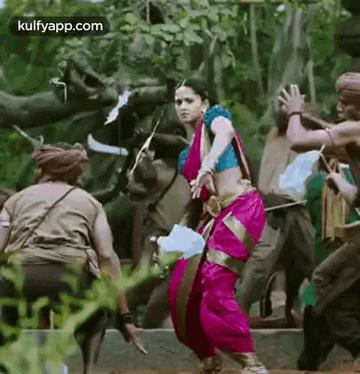 a woman in a pink saree is dancing in front of a group of people in a forest .