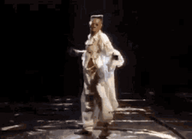 a man in a white robe and gold chains is dancing on a stage with his arms outstretched .