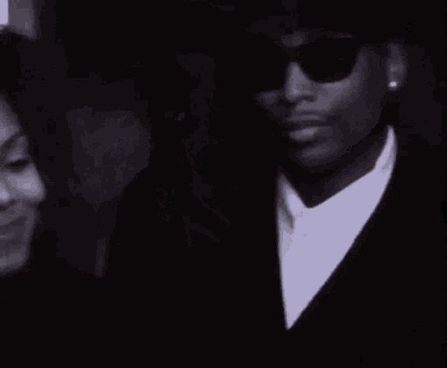 a black and white photo of a man and a woman smiling . the woman is wearing glasses and a hat .