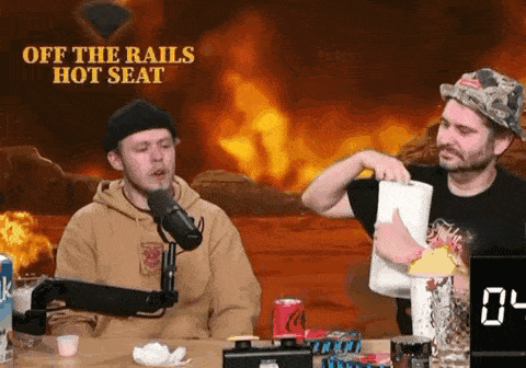 two men are sitting at a table with a microphone in front of a fire and a sign that says off the rails hot seat
