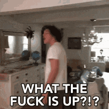 a man in a white shirt is standing in a living room with the words `` what the fuck is up ? ''