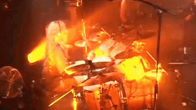 a man playing drums in front of a microphone with a fire behind him