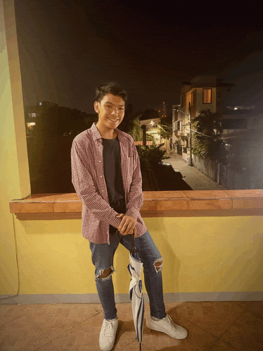 a young man wearing a plaid shirt and ripped jeans stands on a balcony
