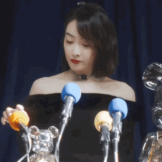 a woman in a black off the shoulder top is standing in front of a bunch of microphones