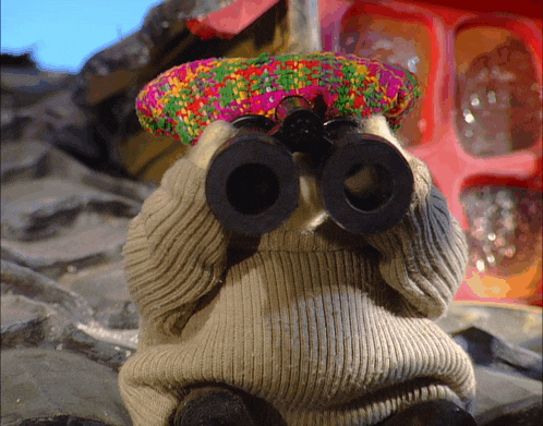 a stuffed animal wearing a colorful hat and sweater looks through binoculars