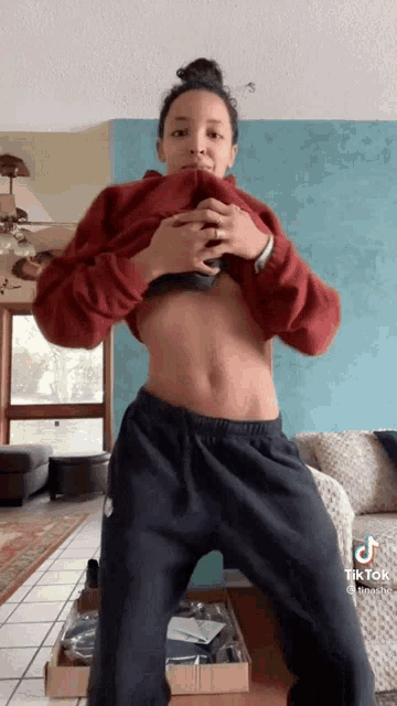 a woman in a red sweater and black sweatpants is standing in a living room with her shirt off .