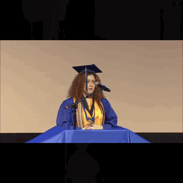 a woman in a graduation cap and gown stands at a podium speaking into a microphone