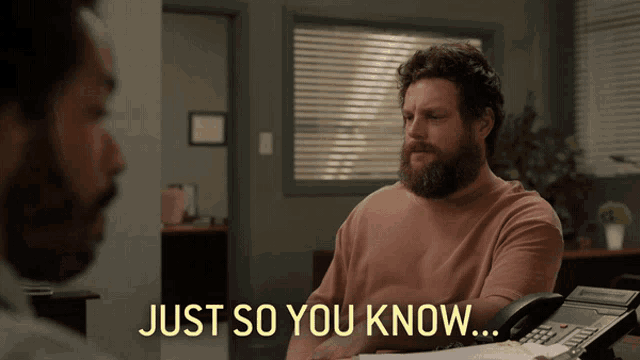 a man with a beard is sitting at a desk with the words just so you know behind him
