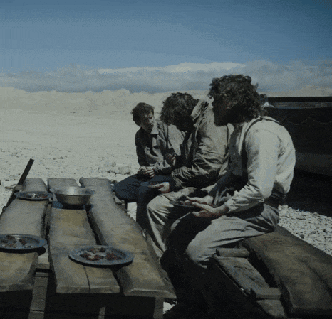 three men sit at a wooden picnic table with plates of food on it
