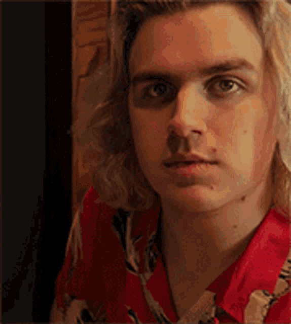 a young man with long blonde hair is wearing a red shirt and looking at the camera