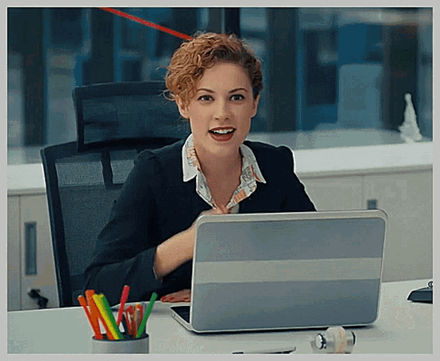 a woman sits at a desk with a laptop and a cup of colored pencils
