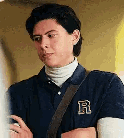 a young man wearing a blue polo shirt and a white turtleneck is standing with his arms crossed and looking at something .