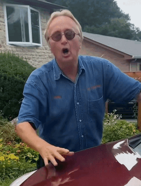 a man wearing sunglasses and a denim shirt stands in front of a brick house
