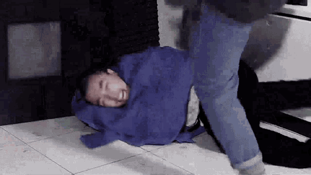 a man is laying on the floor wrapped in a blue blanket while another man kicks him .