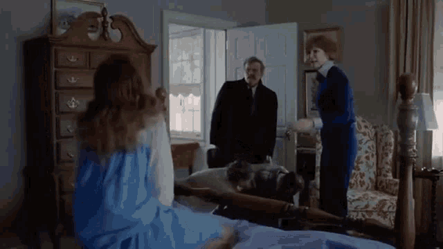 a woman in a blue dress is sitting on a bed in a room while a man in a suit stands nearby .