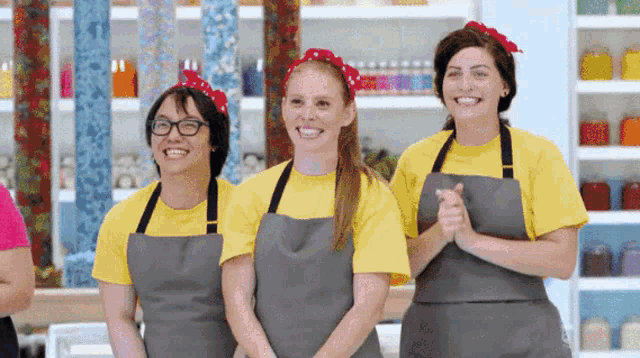 three women wearing aprons and yellow shirts are smiling in a kitchen