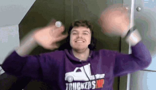 a man wearing a purple hoodie with a truck on it is smiling and waving his hands in the air .
