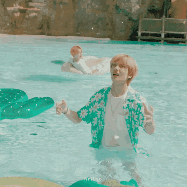 a man in a floral shirt is standing in a pool with another man floating in the background