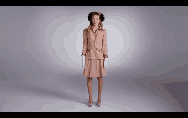a woman in a tan suit and white gloves stands in front of a grey background