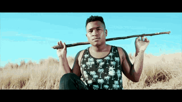 a man wearing a tank top with palm trees on it is holding a stick over his shoulder