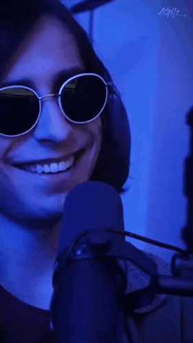 a man wearing round sunglasses is smiling in front of a microphone