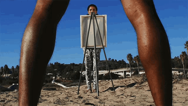 a woman is standing on a beach holding a painting on an easel