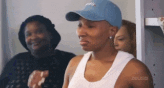 a woman wearing a blue hat and a white tank top is standing in a room with other women .