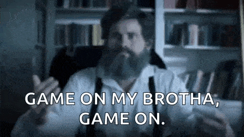 a man with a beard and suspenders is sitting in a chair in front of a bookshelf and talking .