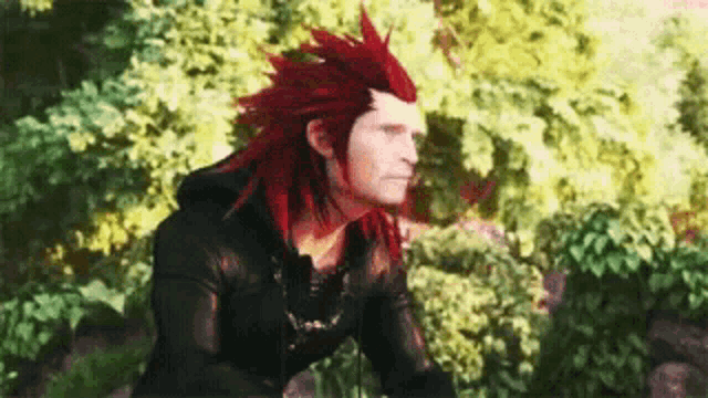 a man with red hair and a black jacket is standing in front of a forest .