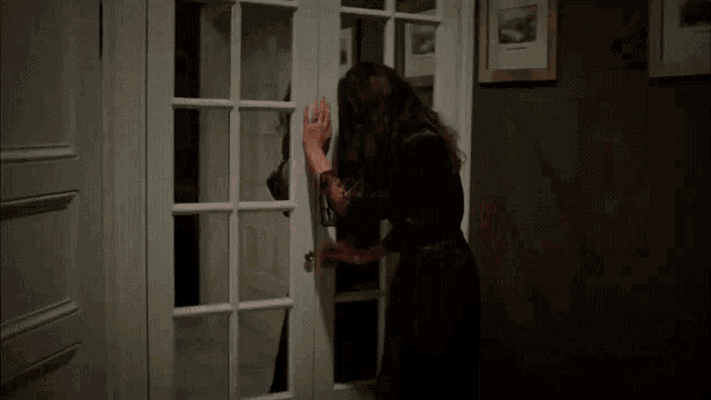 a woman in a black dress is standing in a dark room opening a glass door .