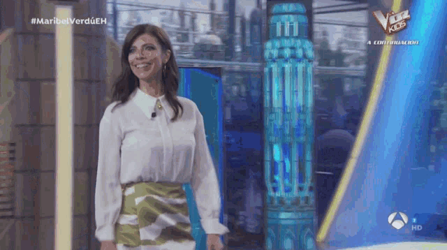 a woman in a white shirt and green skirt is smiling in front of a sign that says the voice kids
