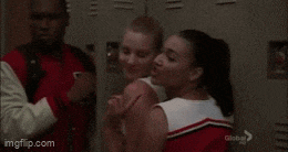 two cheerleaders are standing next to each other in a locker room while a man looks on .