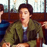 a young man in a green jacket is sitting at a table with a drink and a pen in his hand