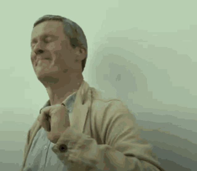 a man in a tan jacket is standing in front of a white wall with his eyes closed and his hands on his chest .